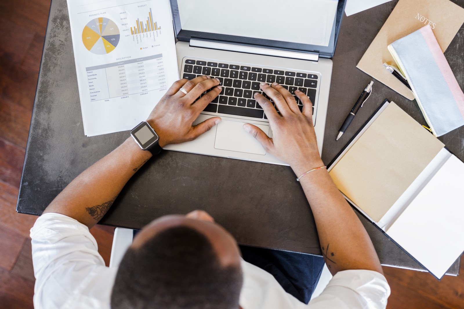 Imagem mostra homem sentado mexendo no notebook com cadernos, papeis com gráficos e outros elementos que remetem a organização financeira