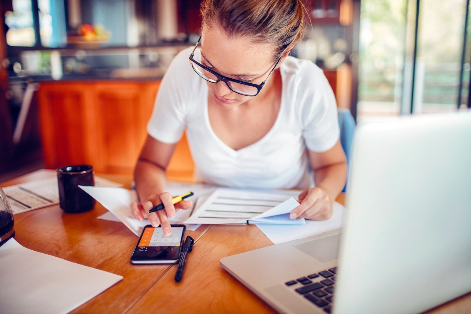 Mulher usa a calculadora de um celular sentada à uma mesa no seu restaurante com o computador aberto e vários papeis