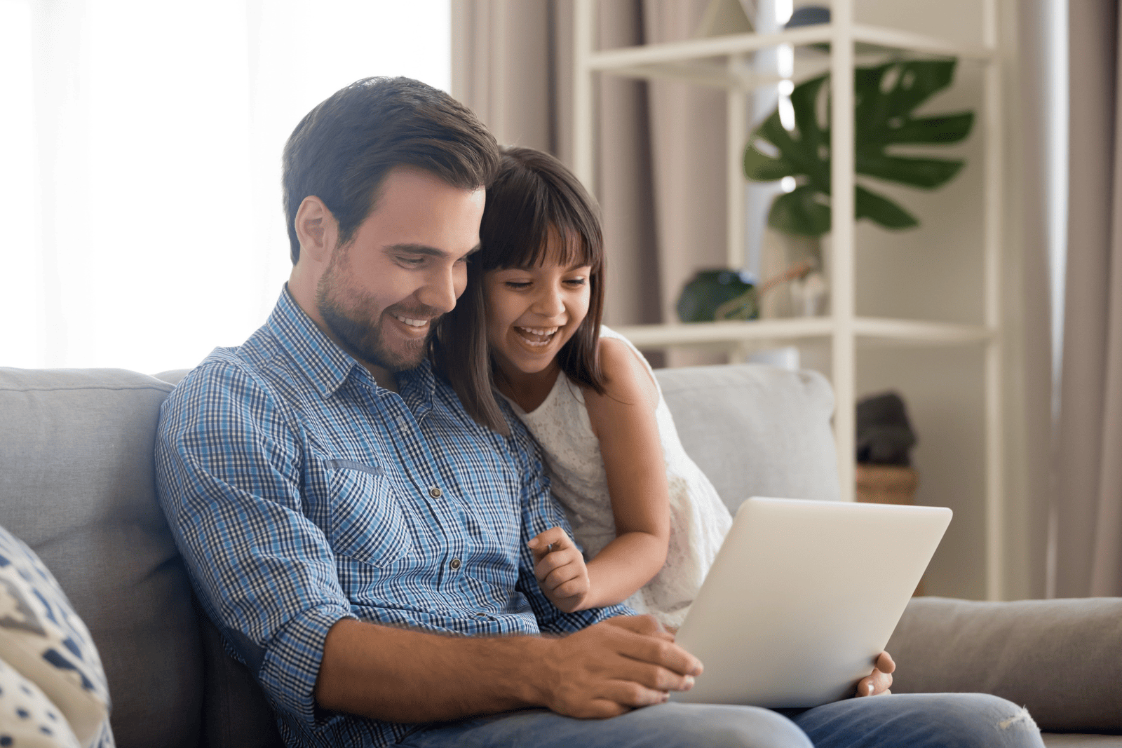 Pai e filha sorrindo enquanto usam um notebook juntos no sofá de casa enquanto fazem compras de Dia dos Pais.
